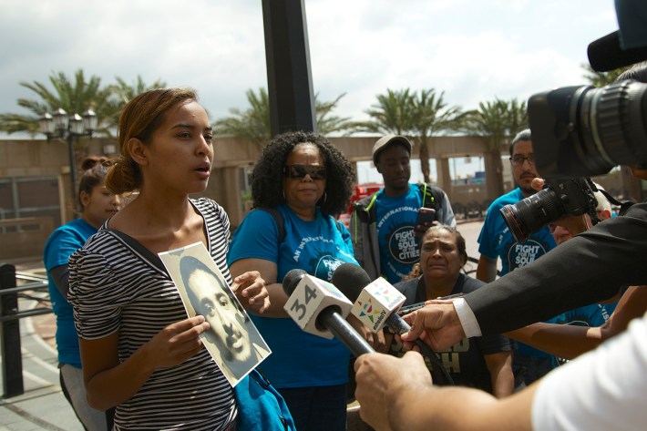 Evelia Granados holds a photo of her brother, Cesar Rodriguez, who was killed by a train while interacting with the Long Beach Police regarding fare evasion. Rodriguez' mother, Rosa Moreno (seated, in the black shirt), looks on as her daughter speaks. Sahra Sulaiman/Streetsblog L.A.