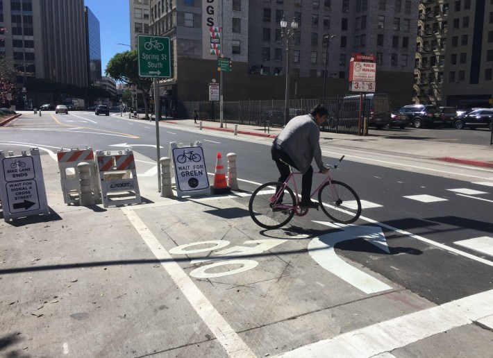 Between 8th and 9th Streets, next to a mid-block signalized crosswalk, cyclists wait in a painted bike box to cross back to trigger a bike crossing signal. Cyclists can continue south in the right-hand bike lane where Spring merges into Main Street.